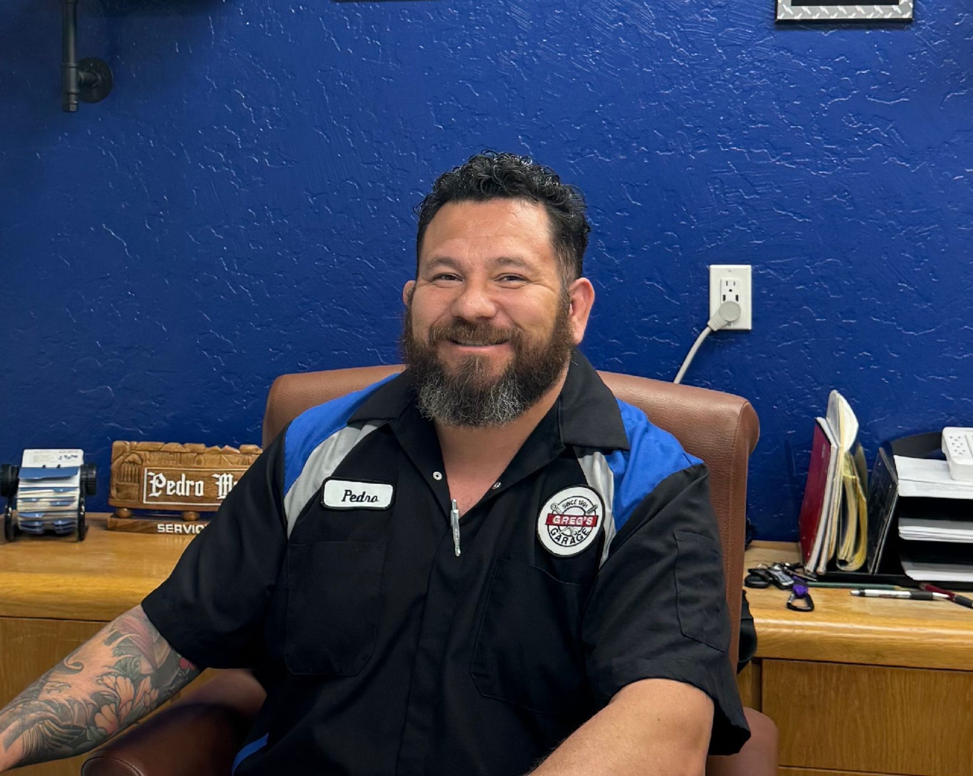 Pedro Mendoza - Man with beard in auto shop uniform, sitting at desk, smiling. | Greg's Garage