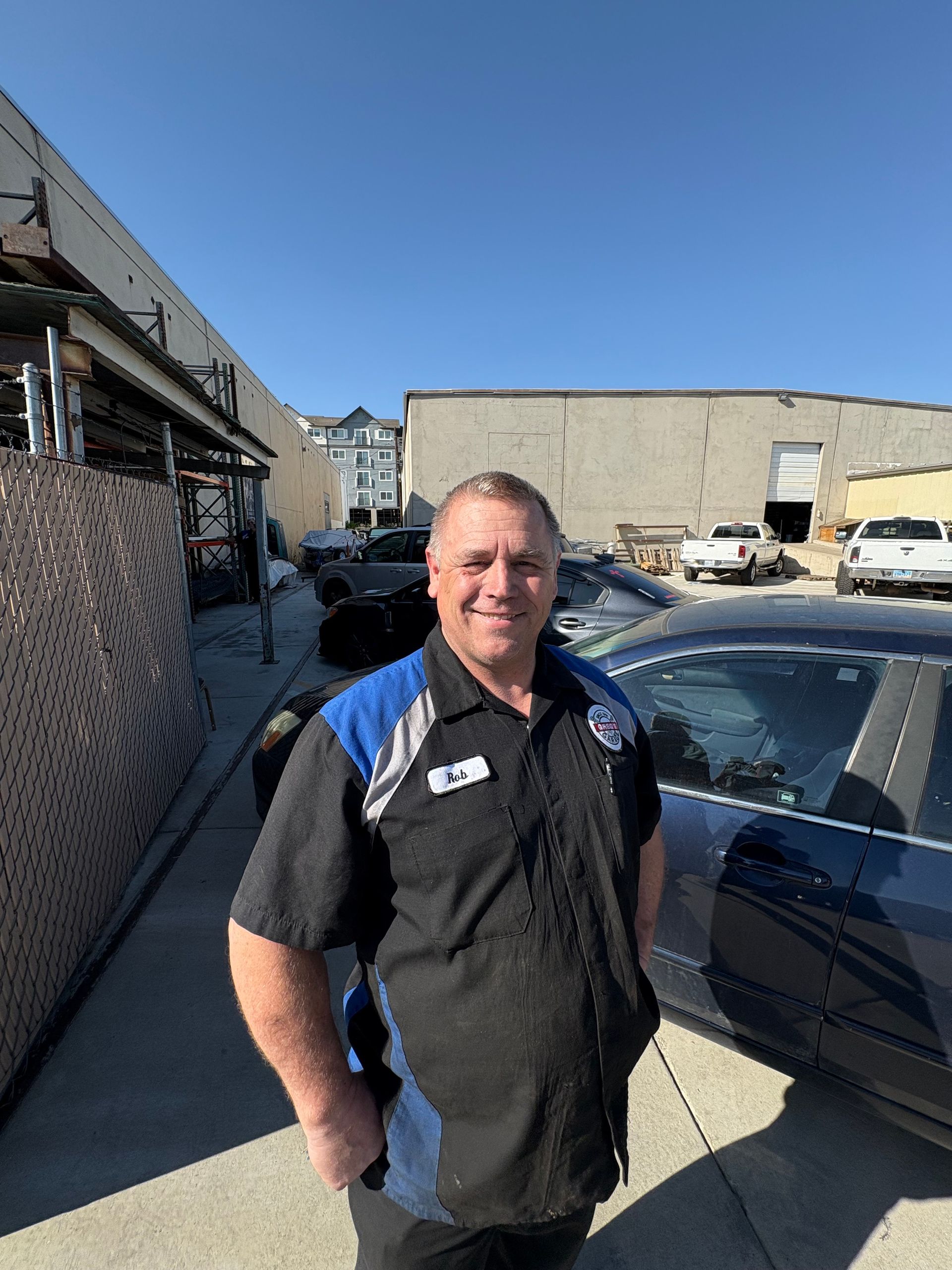 Man in mechanic uniform standing outdoors with cars in the background. Blue sky. | Greg's Garage