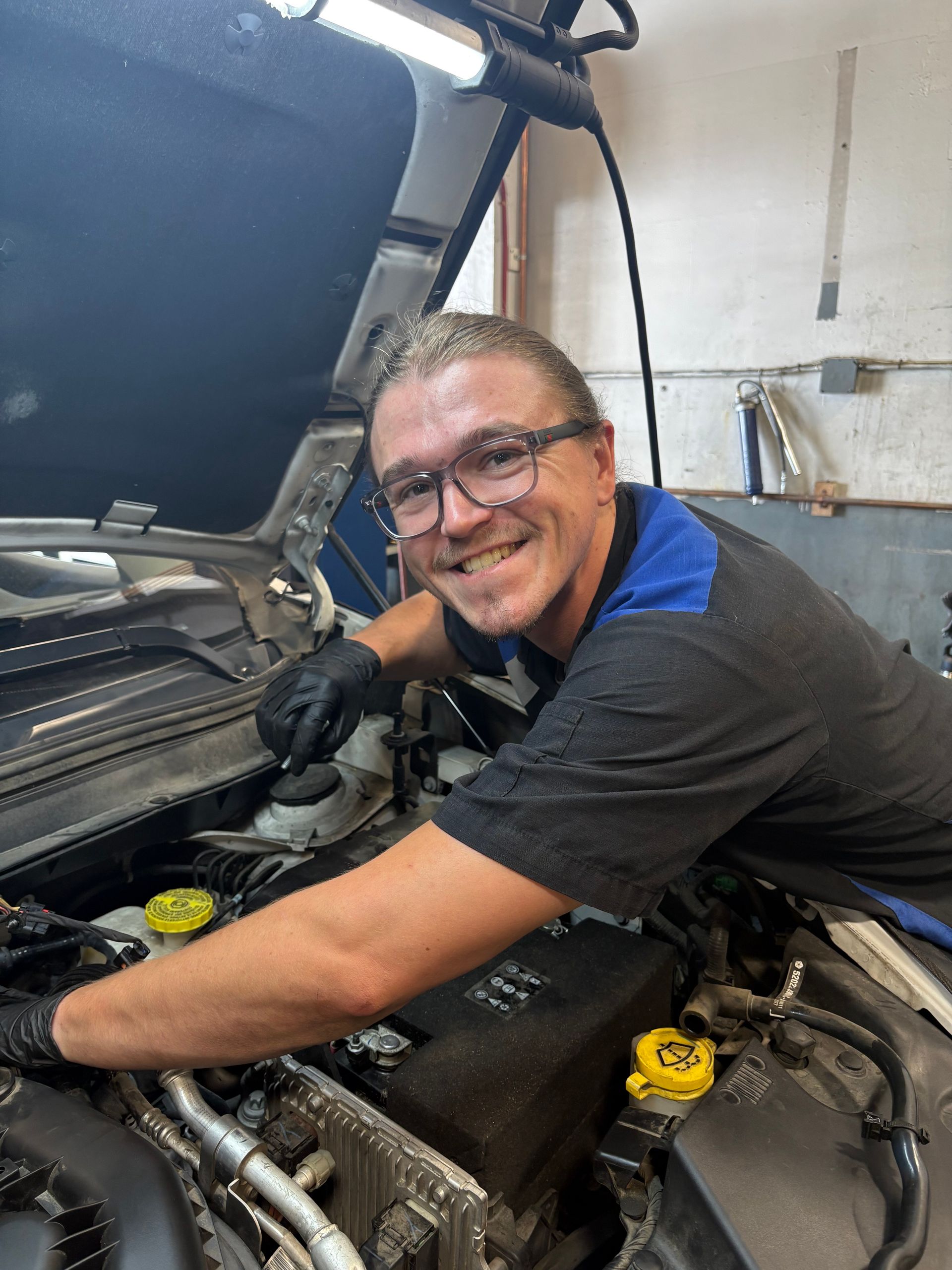 Mechanic working on car engine, smiling at camera. Black gloves, glasses, dark shirt. Garage setting. | Greg's Garage