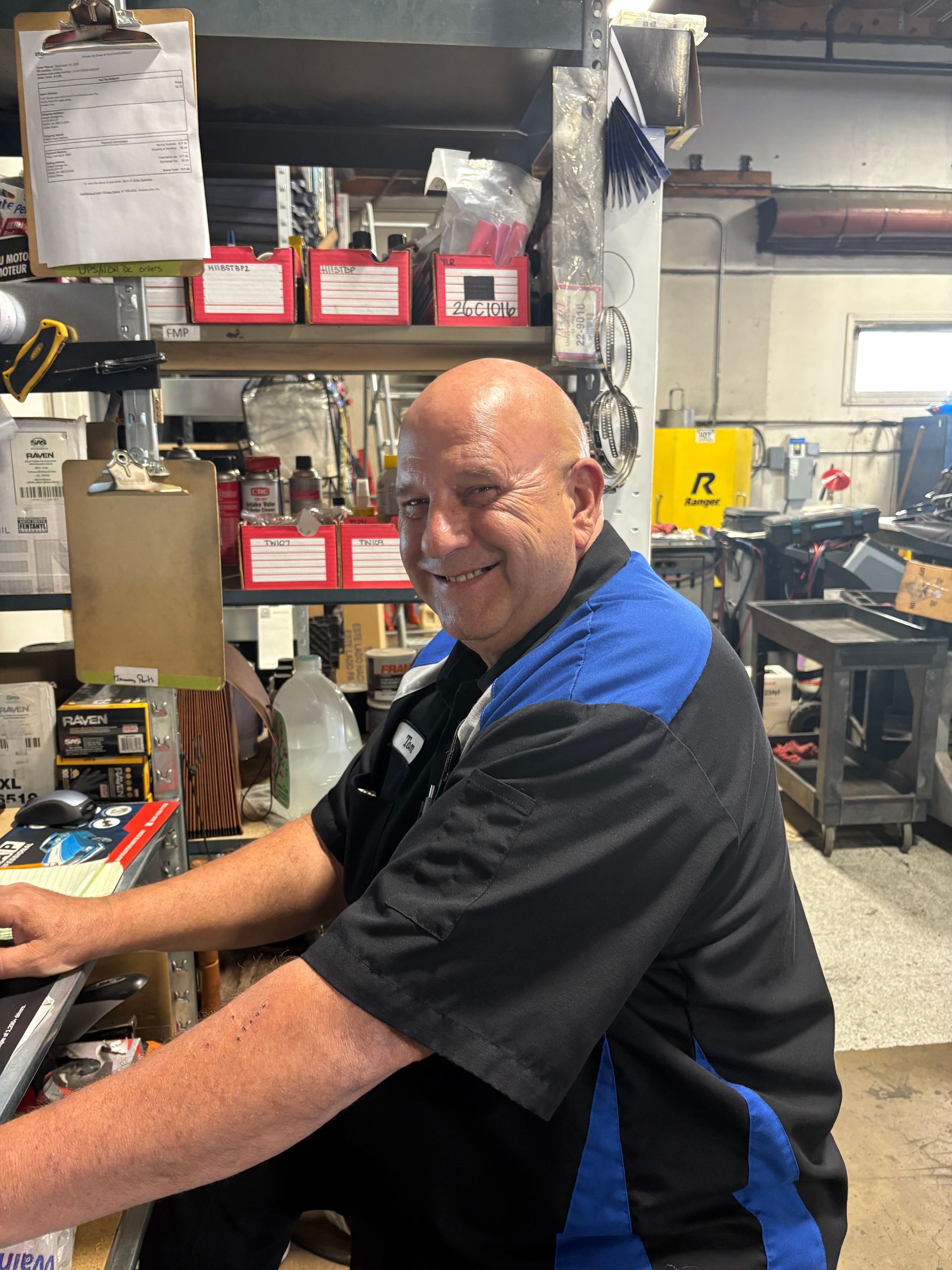 Man in mechanic shop, wearing black and blue shirt, smiles at camera while working at a computer. | Greg's Garage