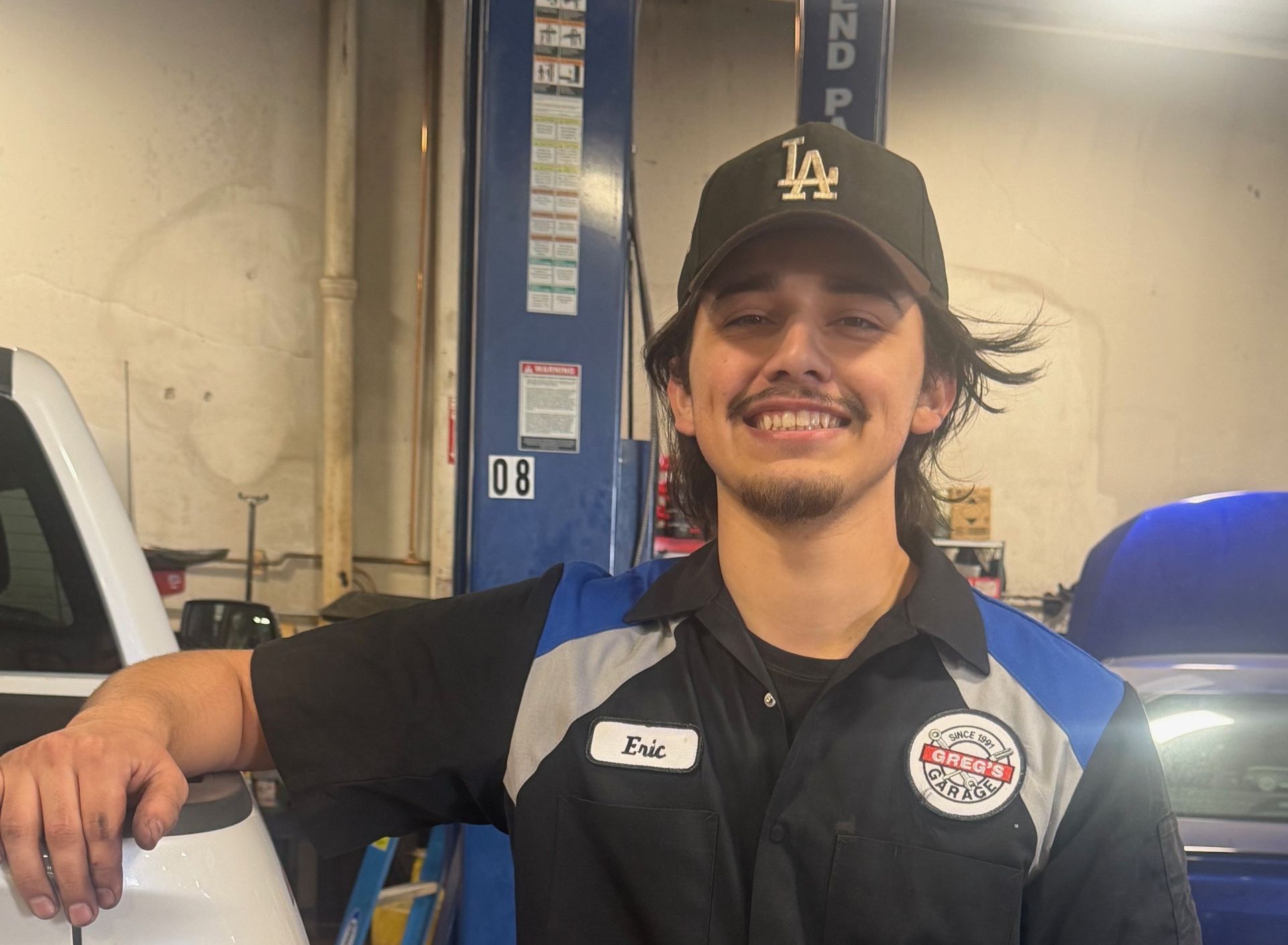 Eric - a smiling mechanic wearing Greg's Garage uniform and a black baseball cap posing next to a vehicle lift.