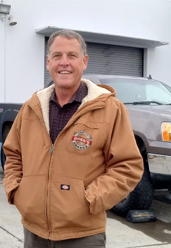 Greg Doyle, Owner, in brown coat smiling, standing in front of a building and a truck. | Greg's Garage