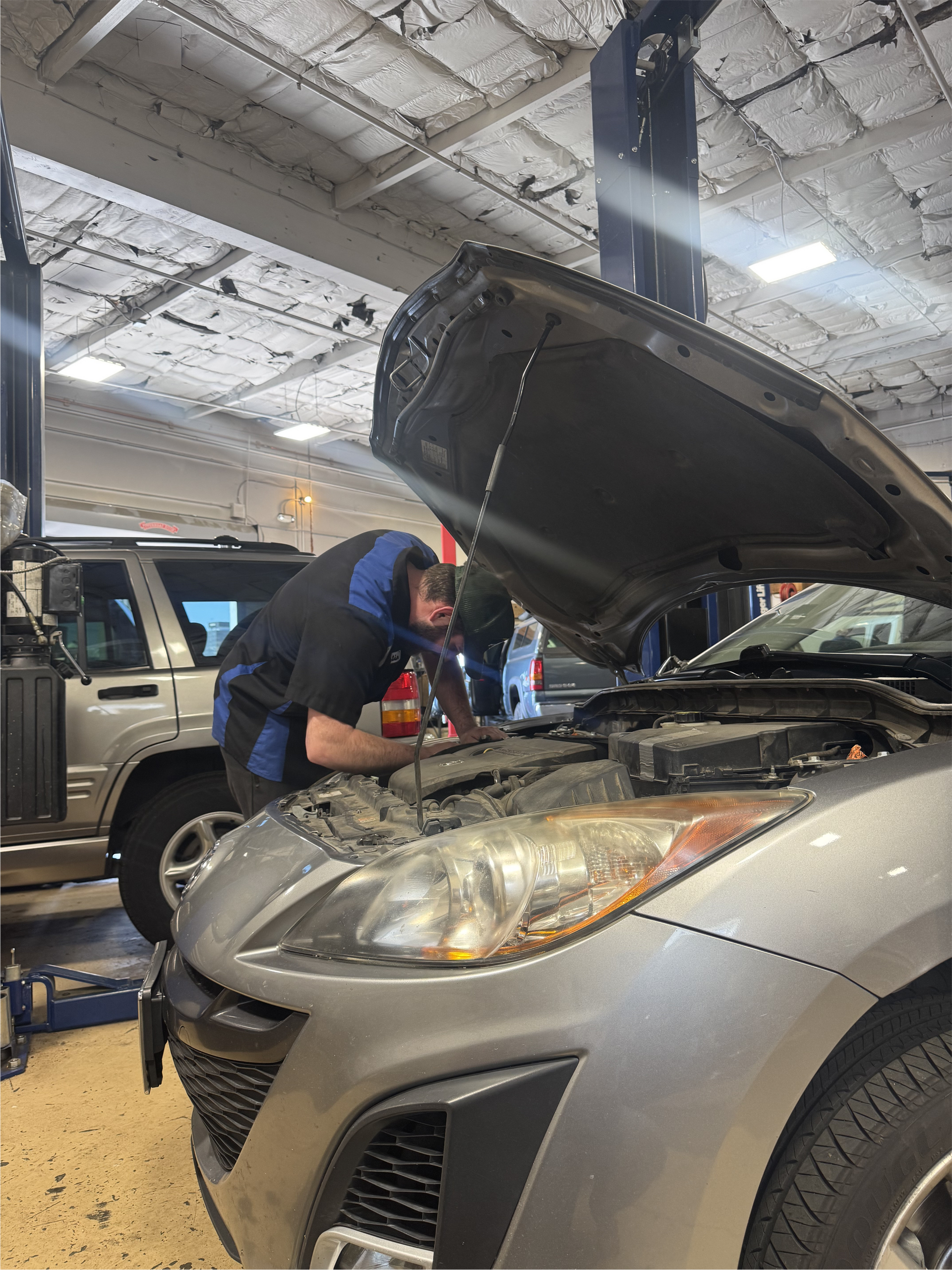 Mechanic working on a silver car in a garage. Hood is open. Another vehicle in the background. | Greg's Garage