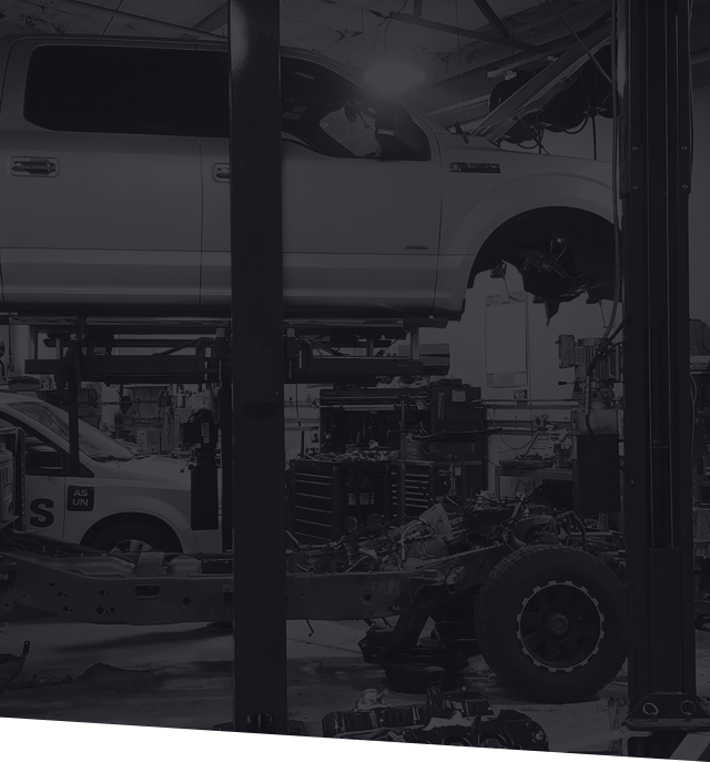Truck on a lift in a mechanic's shop. Parts, tools, and a second vehicle are visible in the grayscale photo. | Greg's Garage