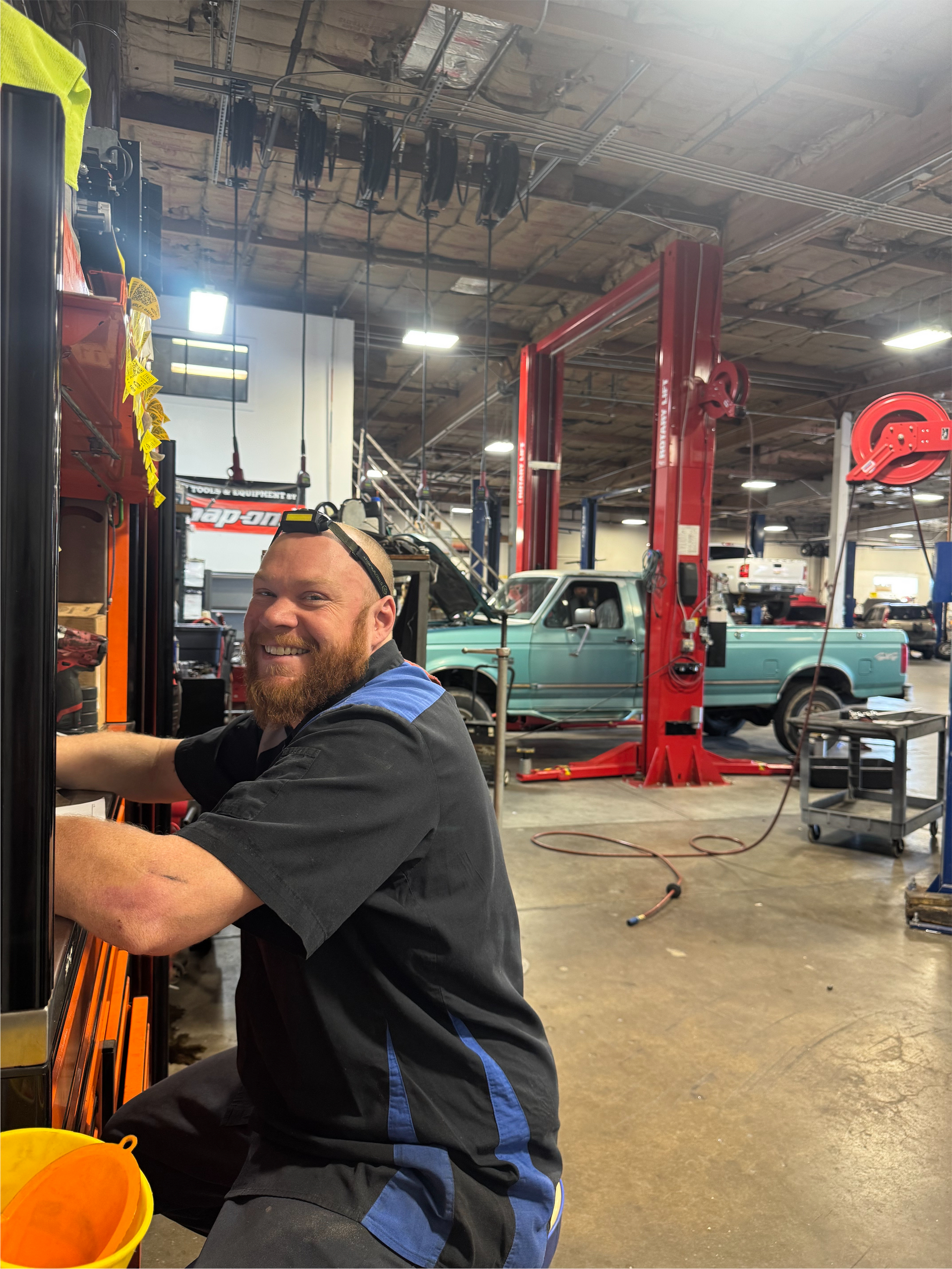 Mechanic in a garage, smiling, working on equipment with a light blue truck in the background. | Greg's Garage