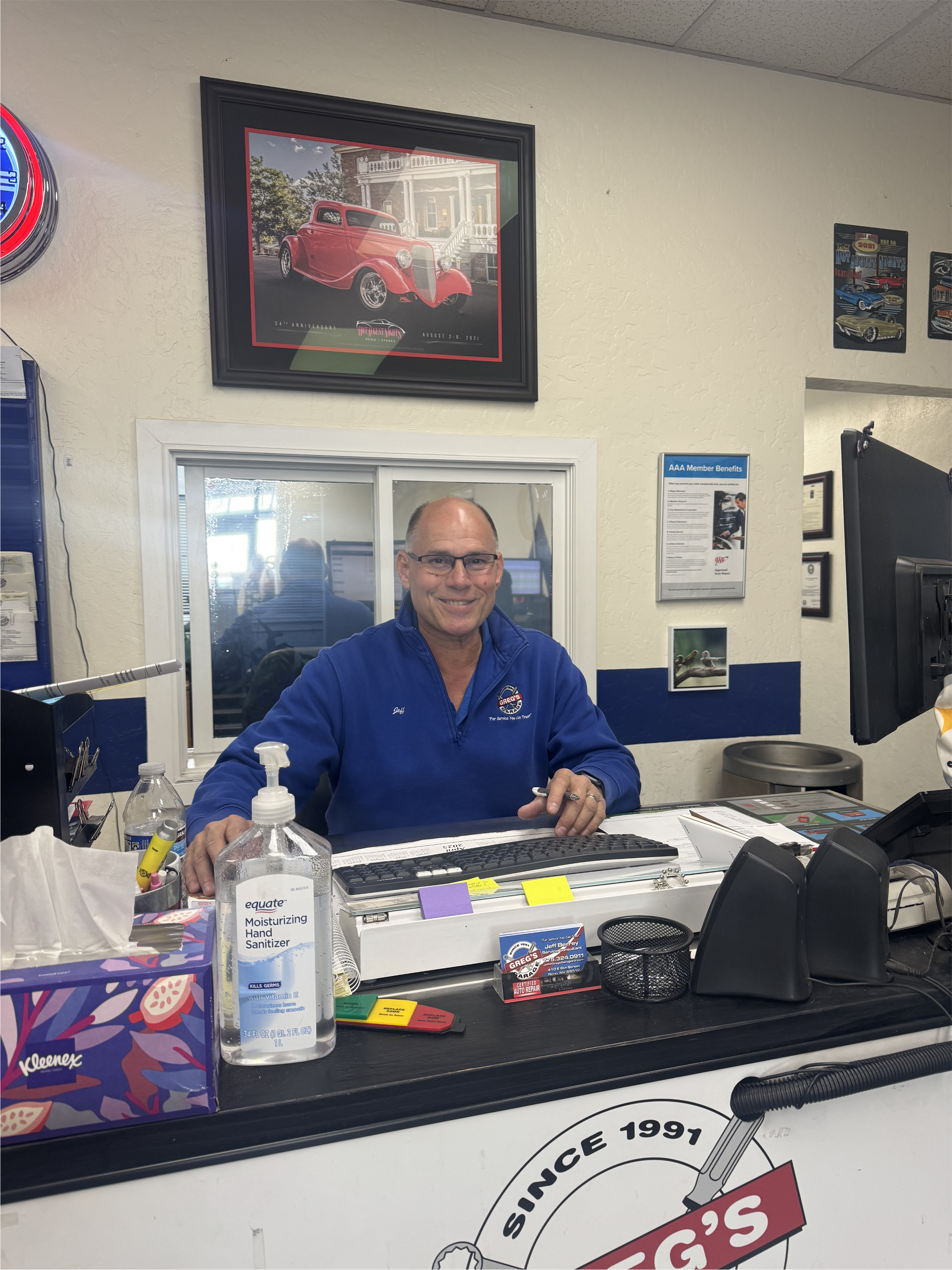 Man at a car repair shop counter smiles, wearing blue. Shop interior has a framed photo and hand sanitizer. | Greg's Garage