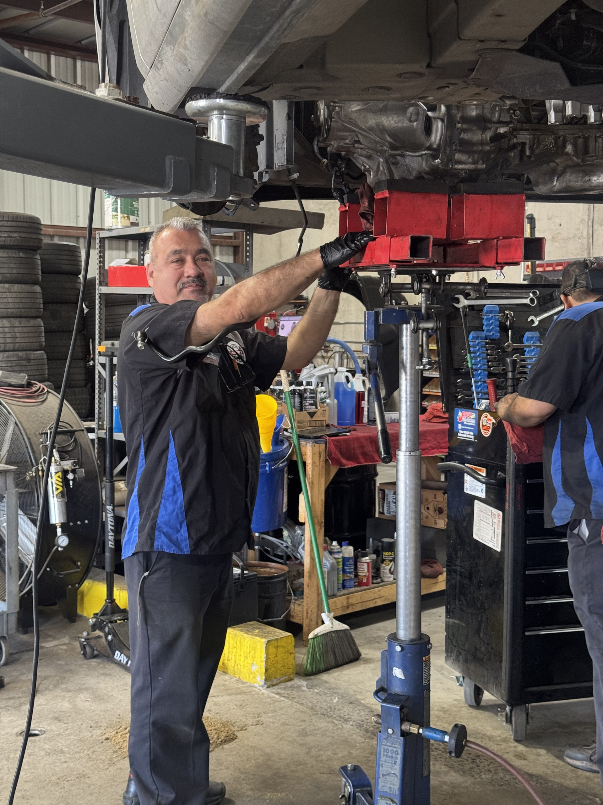 Mechanic working on a car lift in a shop. Wearing blue and black shirt, smiling, and pointing. | Greg's Garage