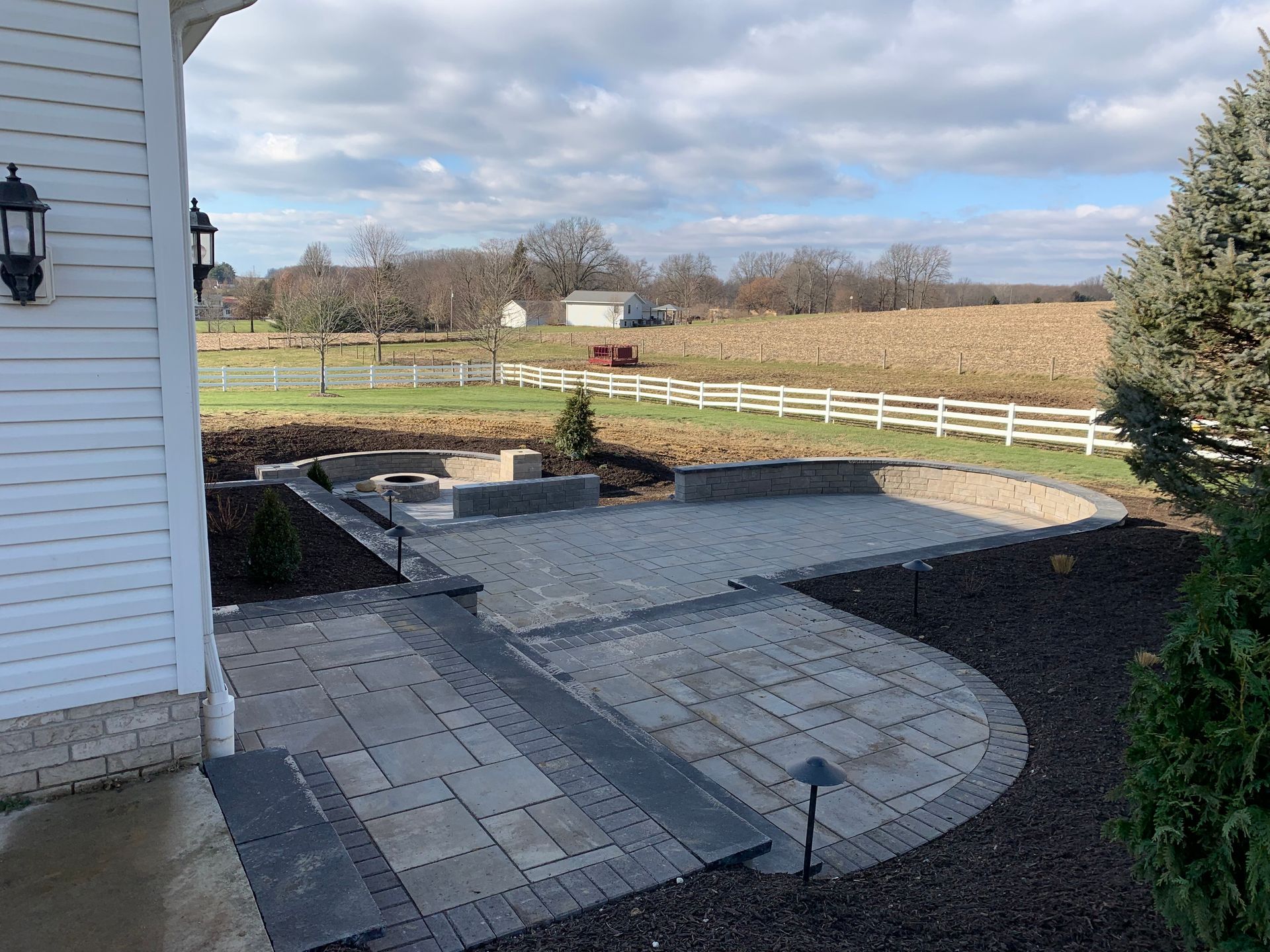 Stone patio with steps and landscaping, overlooking a field with a white fence under a cloudy sky.