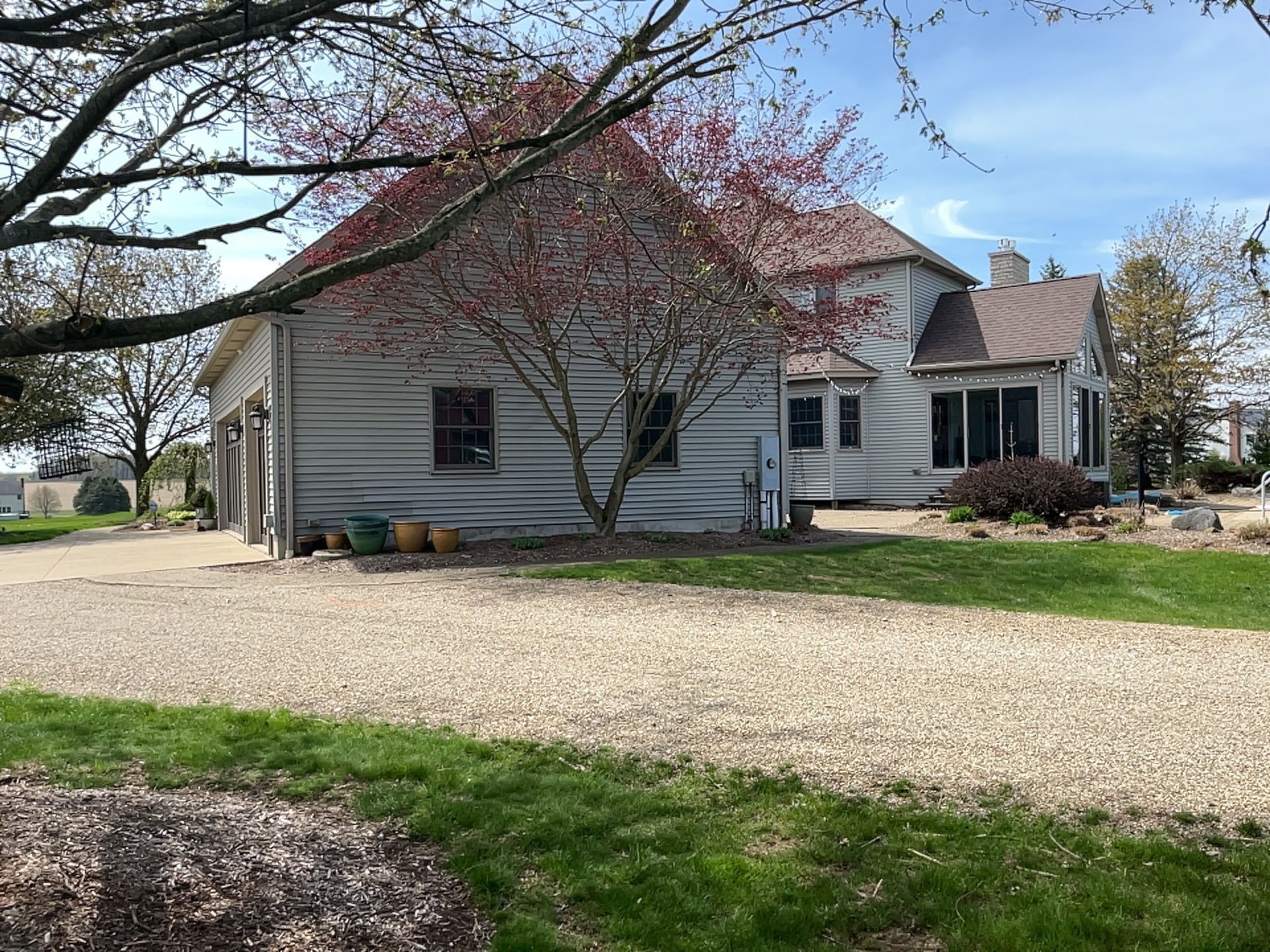 Gravel driveway leading to a weathered, two-story house with attached garage; pink-flowering tree in front.