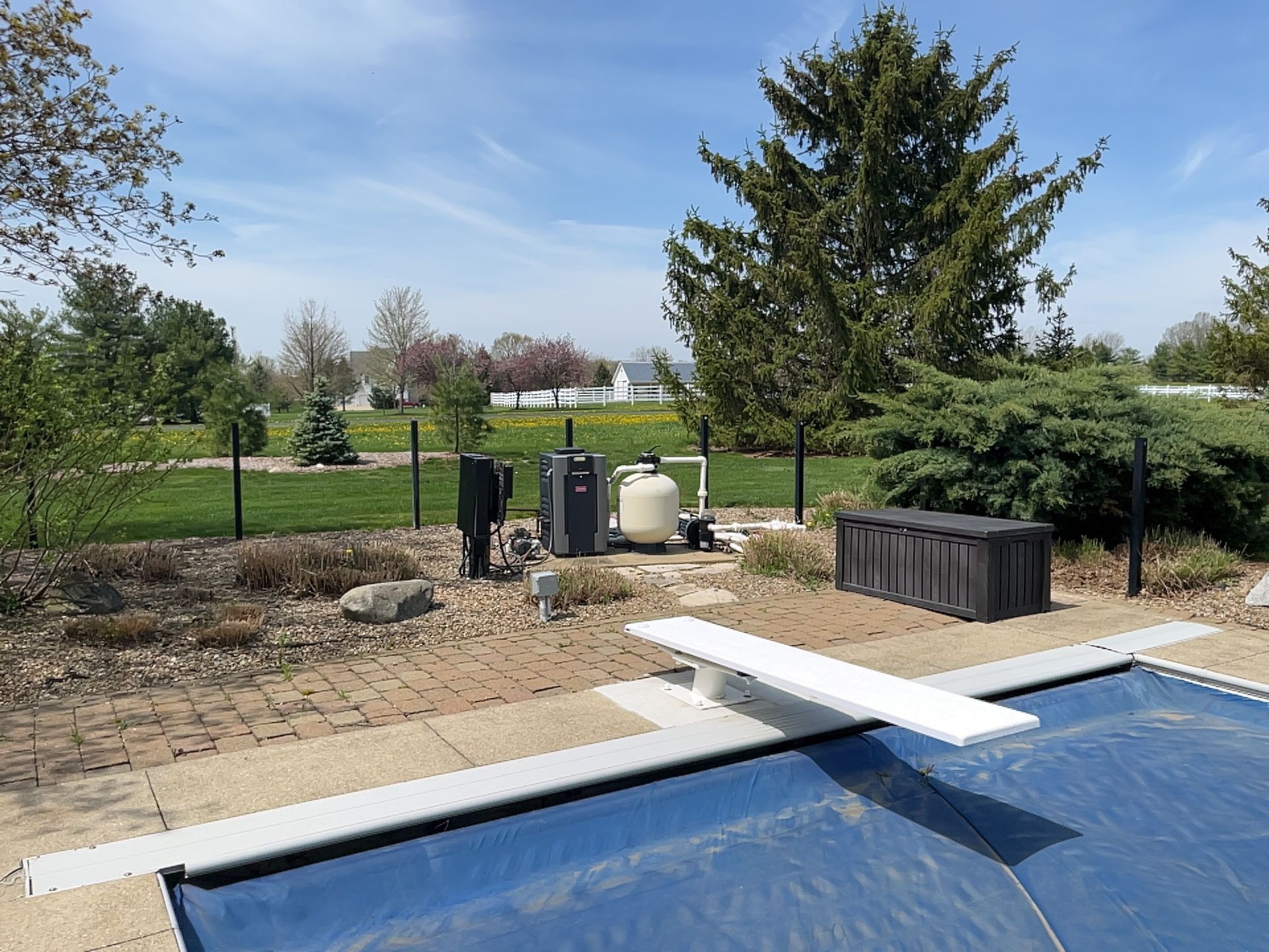 Poolside view with diving board, equipment, and blue pool cover, under a blue sky.