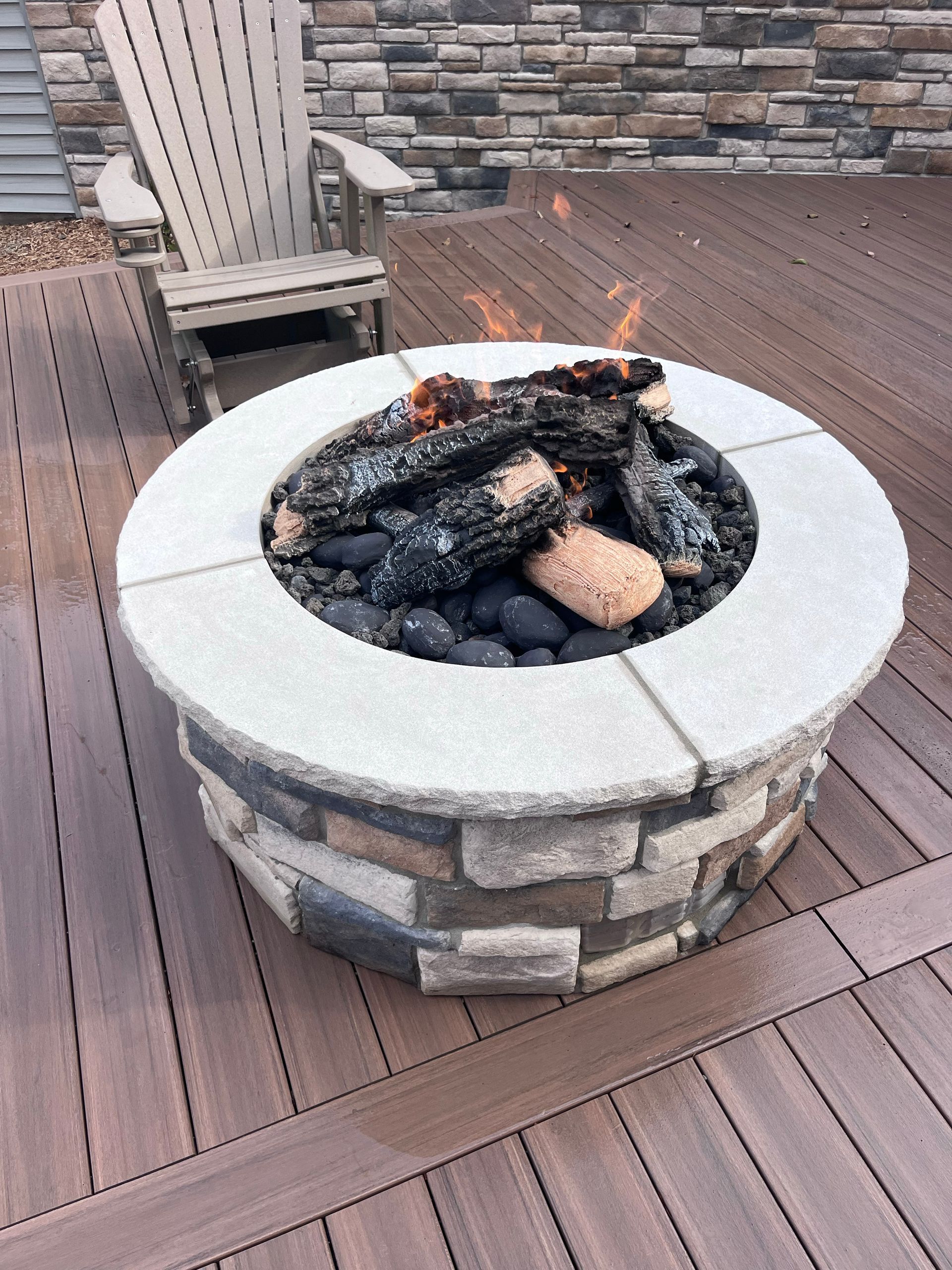 Stone fire pit with burning logs on a wooden deck next to a wooden chair.