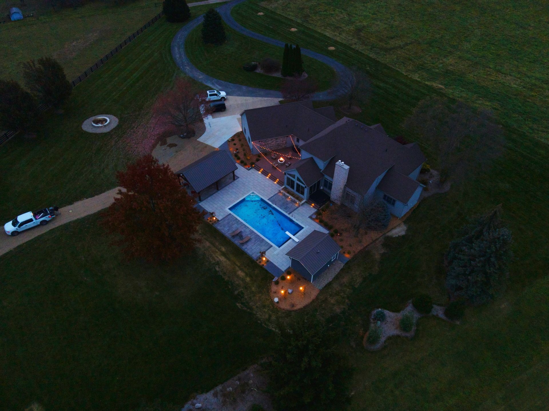 Aerial view of a house with a pool, driveway, and surrounding green lawn in a rural setting at dusk.