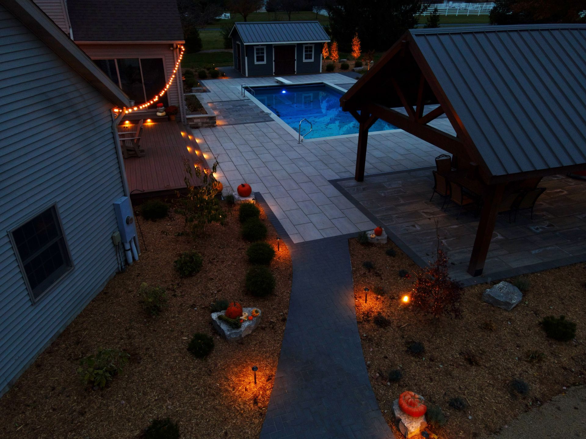 Nighttime backyard scene with lit pool, patio, and path lined with pumpkins and lights.