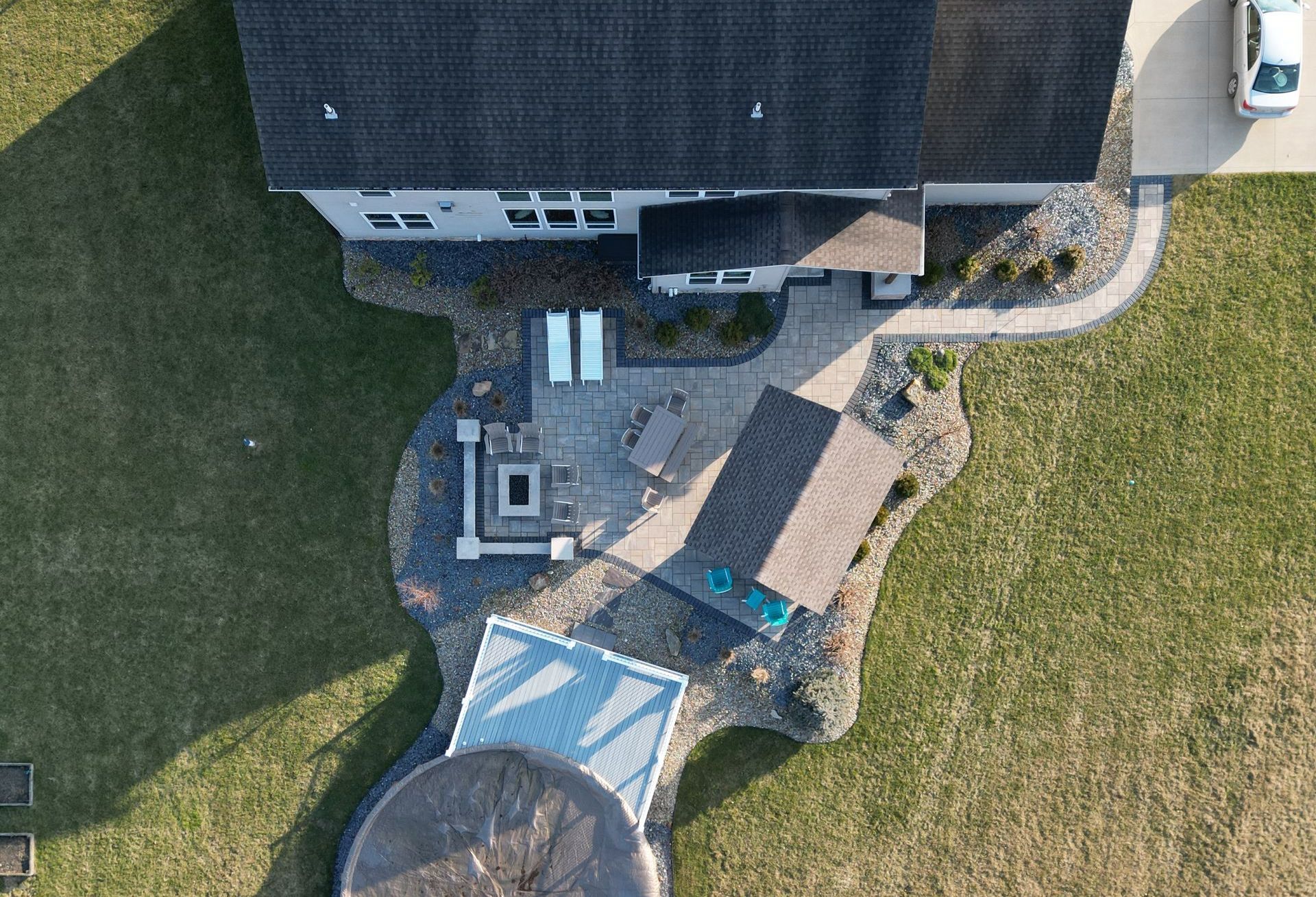 Aerial view of a backyard patio with a fire pit, seating, and a pergola; pathway leads to the driveway.