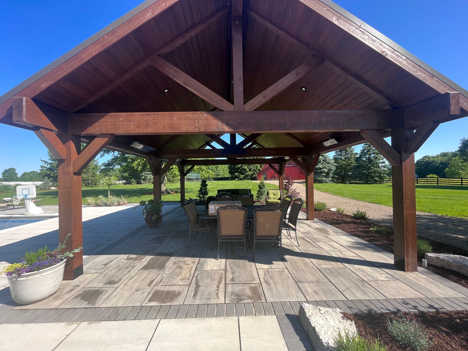 Brown wooden gazebo over patio with dining table and chairs, on a sunny day.