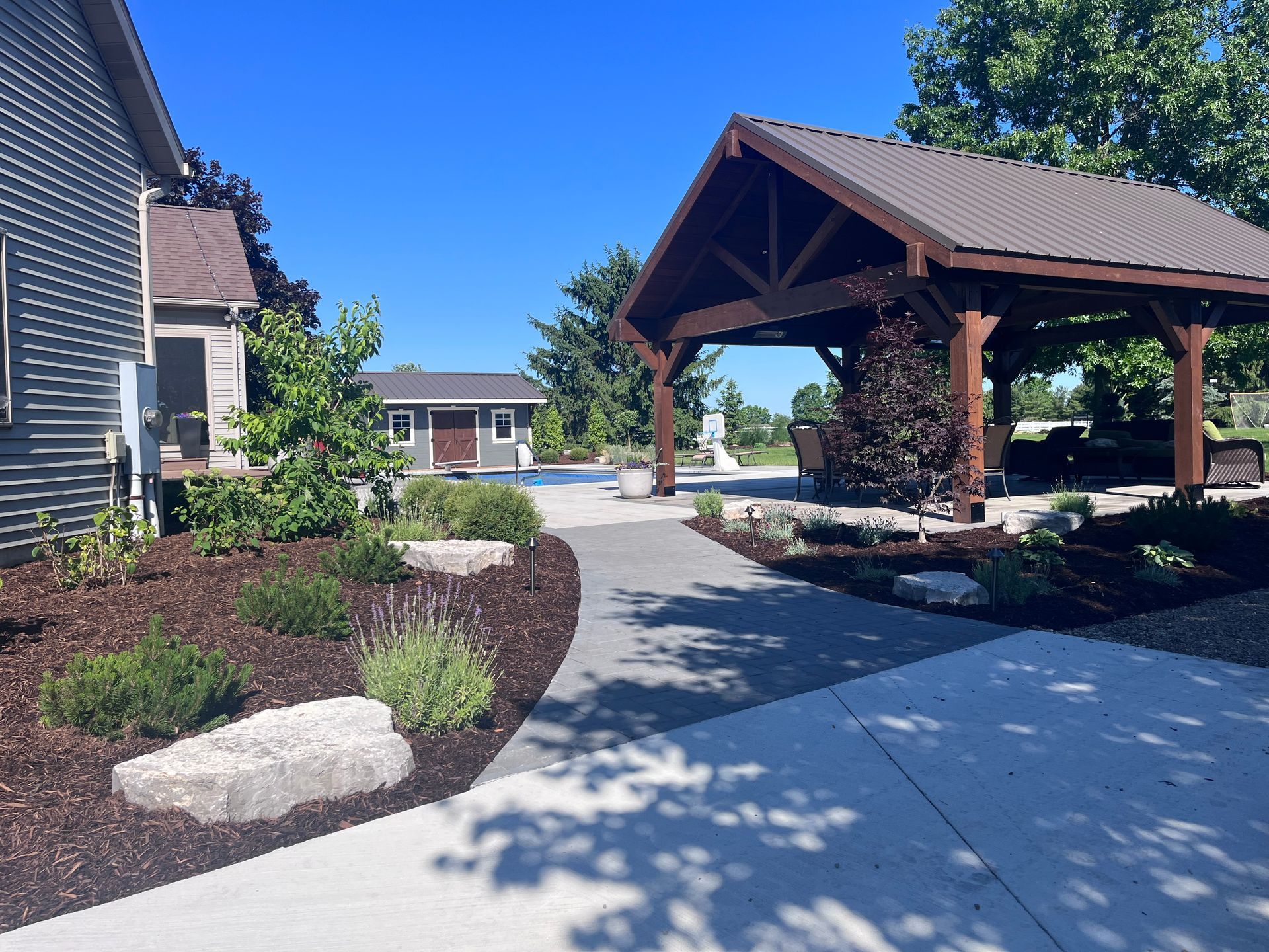 A concrete walkway leads to a wooden gazebo. A building is on the left, and a small structure is in the distance.