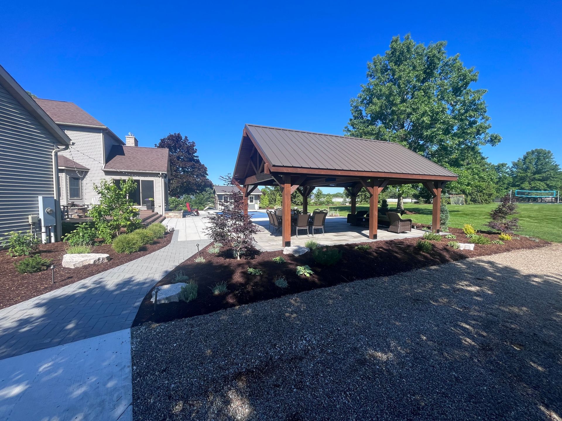Patio with gazebo, seating area, house, landscaping, gravel driveway, and blue sky.