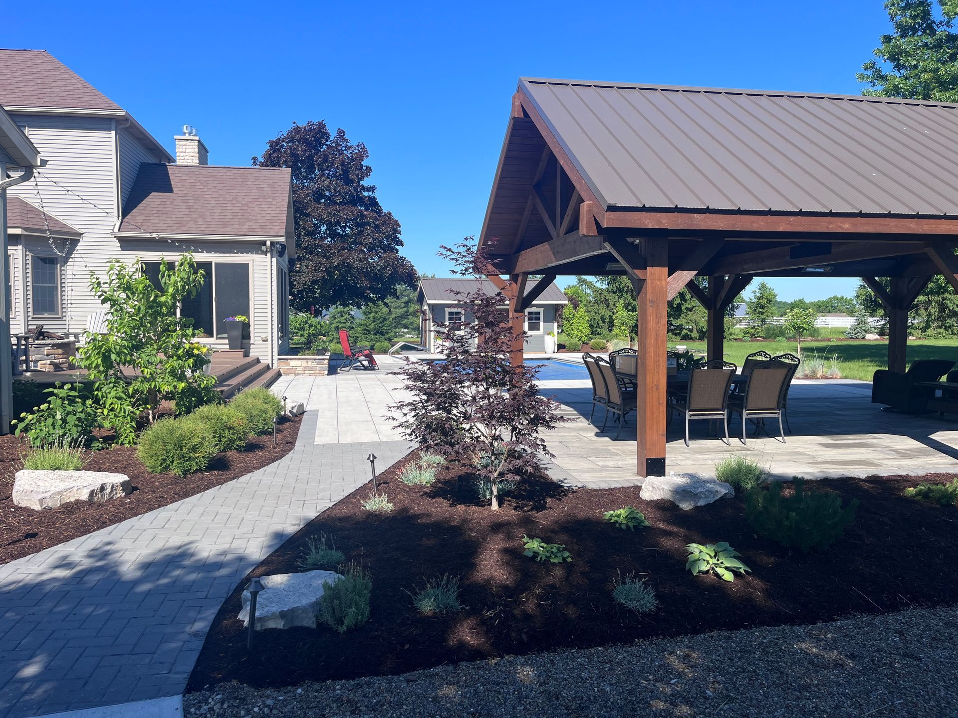 Patio with a pavilion, dining table, and seating by a house with landscaping on a sunny day.