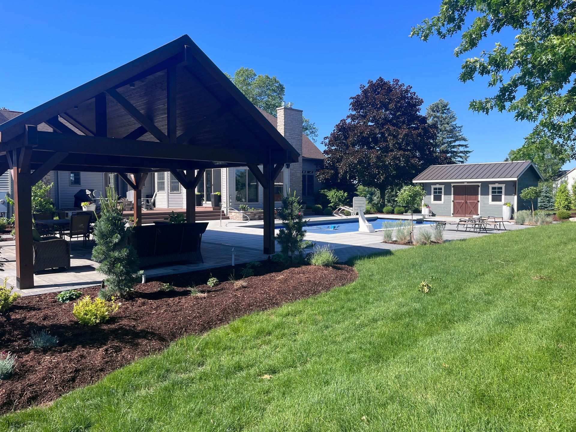 Backyard with a dark wooden pergola, pool, small shed, and green lawn on a sunny day.