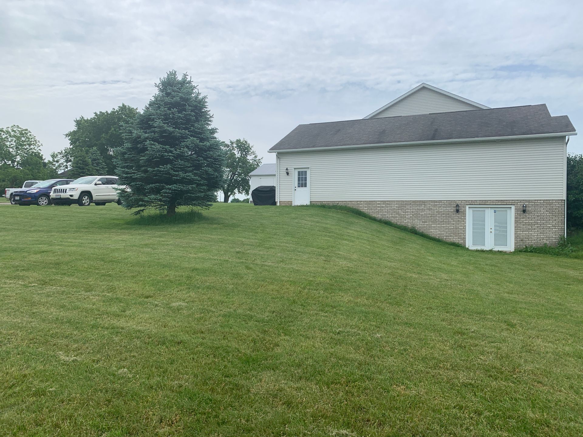 Green lawn with a two-story building on a small hill; a shed and cars are visible. Overcast sky.