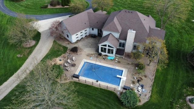 Aerial view of a large house with a swimming pool, driveway, and surrounding green lawn.