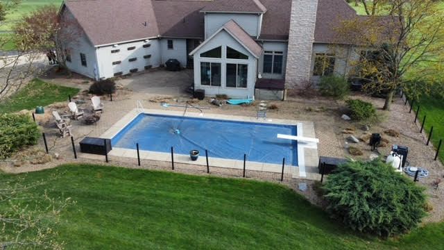 Backyard with a swimming pool covered by a blue tarp, beside a house with tan siding and a brown roof.