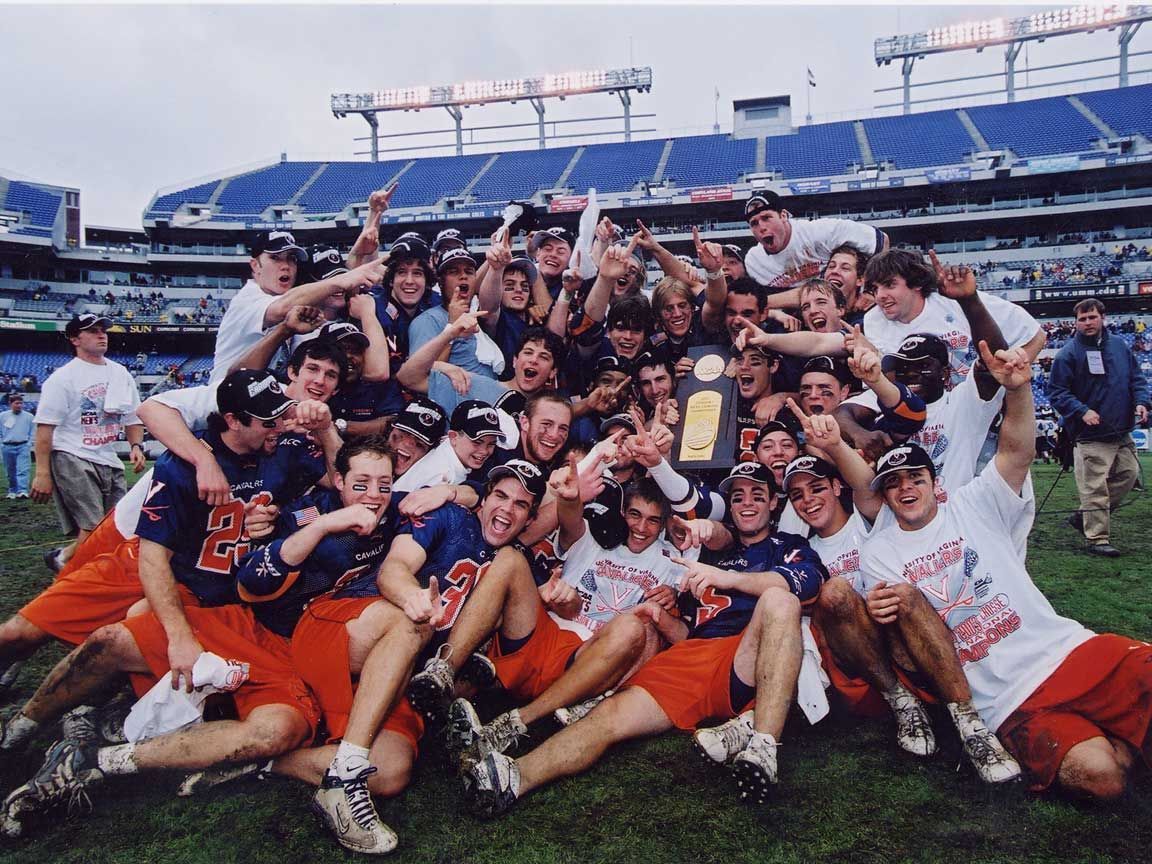 2003 National Champions University of Virginia (UVA) Lacrosse team celebrates championship on field in Baltimore at M&T Bank stadium. Players wear orange, navy blue, and white; smile, cheer, hold trophy.