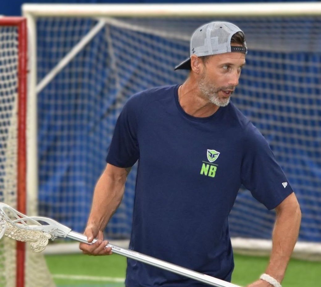 Ned Bowen coaching Fusion lacrosse holding a lacrosse stick, wearing a cap and dark blue shirt, with a net in the background.