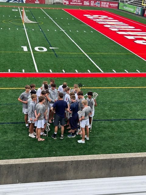 FuturesLC 2029 Lacrosse team huddles on Stoney Brook University field before the 2025 NLF National Championship. Players in white shorts and gray shirts listen to coach Ned Bowen near the red sideline.