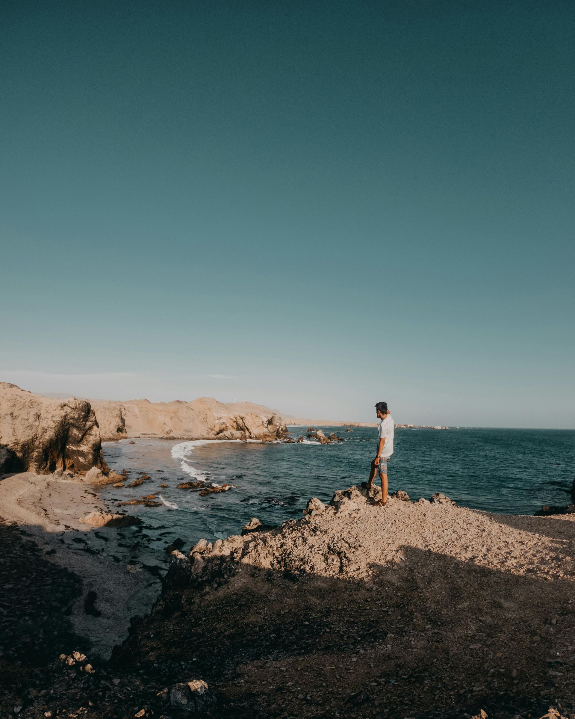 Person standing on a rocky shore, looking out at the ocean under a clear blue sky.
