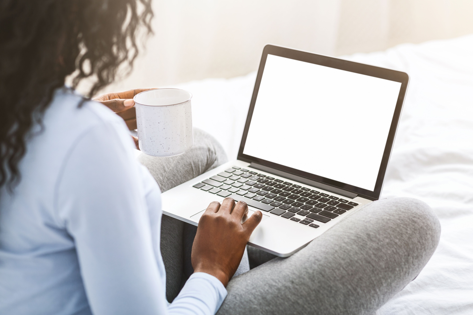 A person in a light blue shirt sits on a white bed with a coffee mug, using a laptop with a blank white screen.