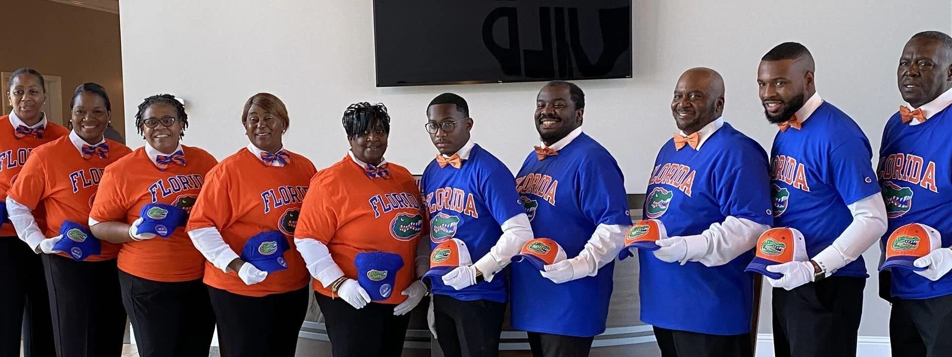 A group of people wearing blue or orange shirts and white gloves, holding matching hats in an indoor setting.
