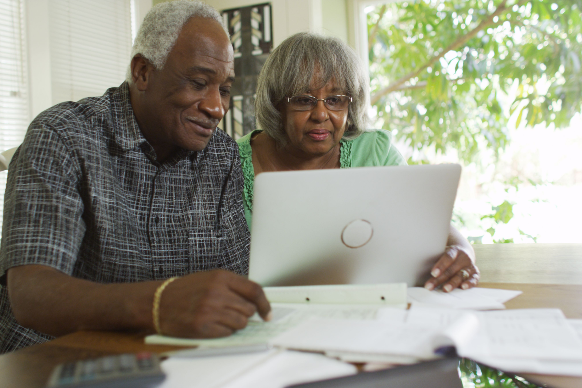 An elderly couple is sitting at a table with a laptop and a notebook.