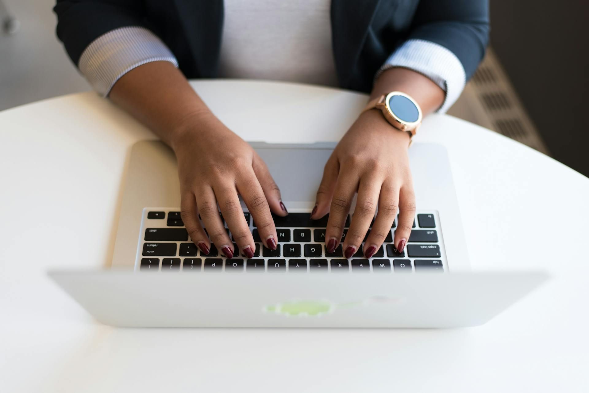 A person wearing a watch typing on a silver laptop on a white desk.
