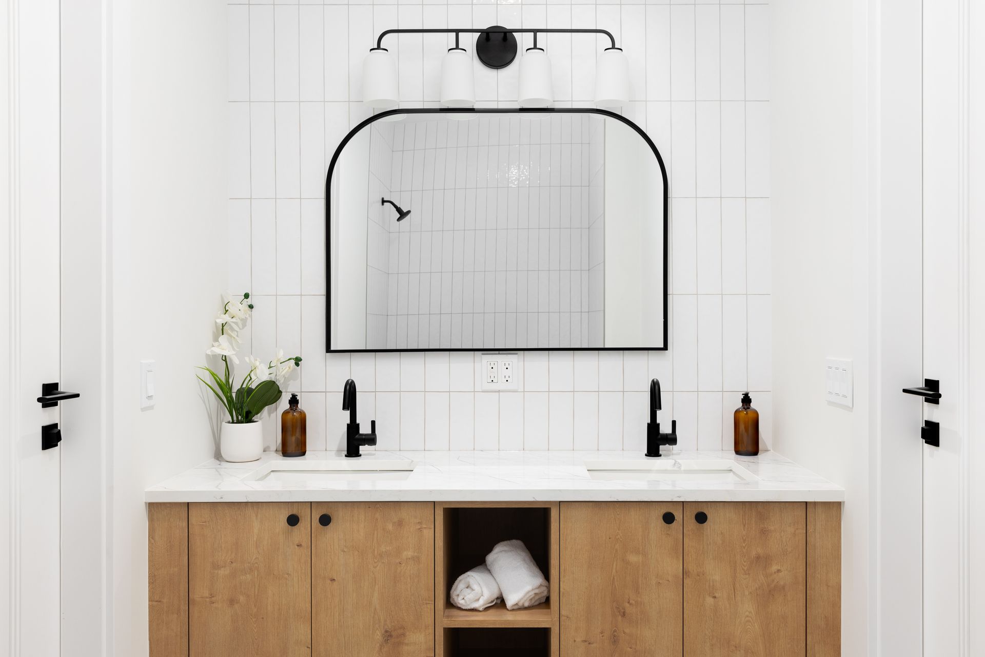 Meuble-lavabo moderne à double vasque avec armoires en bois, robinetterie noire et grand miroir arqué sur des murs carrelés blancs.