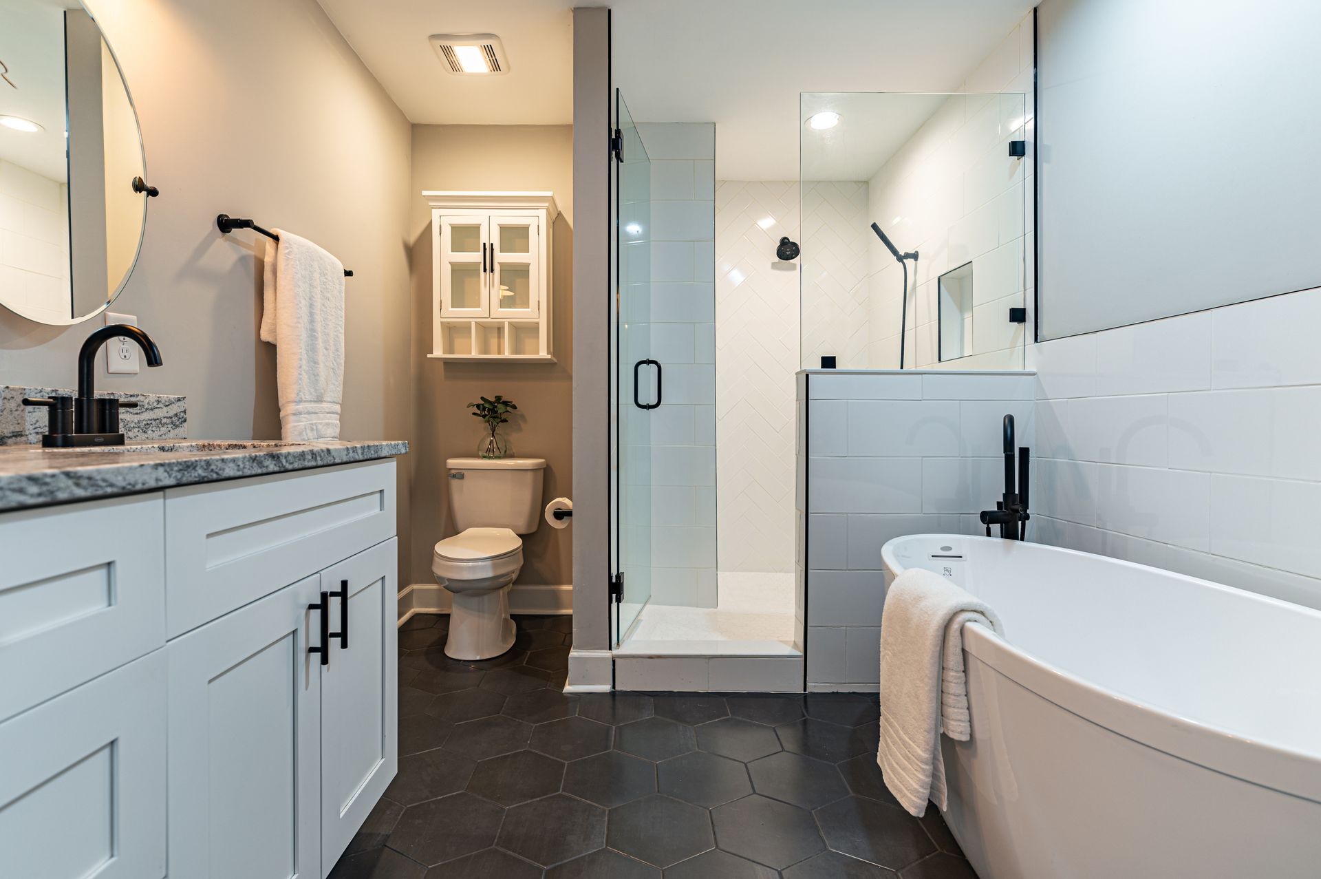 Salle de bains moderne avec meubles blancs, baignoire, douche et sol en carrelage hexagonal noir.
