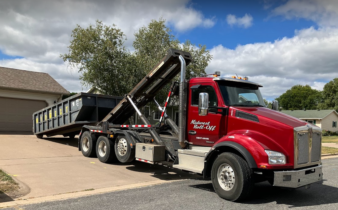 A bright red roll-off dump truck dropping a large gray metal dumpster onto a residential driveway on a sunny day.