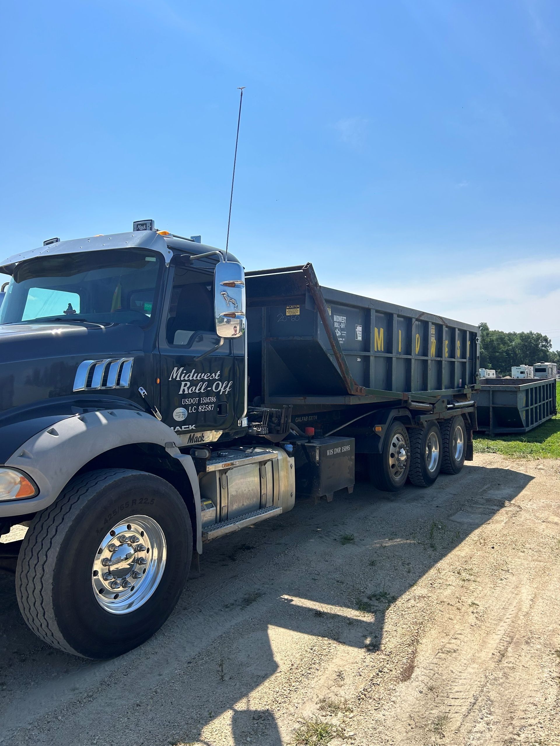 A black roll-off dump truck parked on a gravel lot under a clear blue sky.