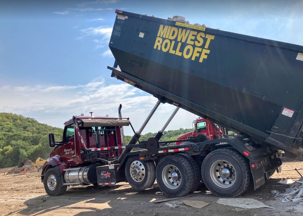 A maroon roll-off truck dumps a large black container at a rural outdoor site under a sunny sky.