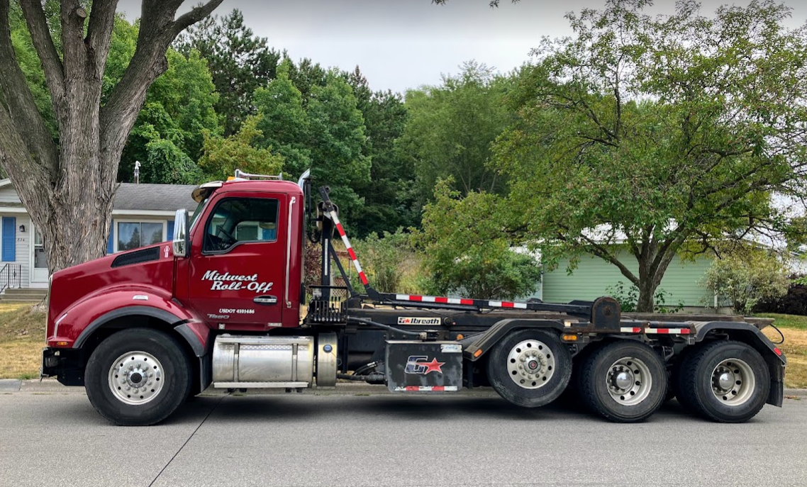 A side view of a maroon roll-off dump truck parked on a residential street with trees and houses in the background.