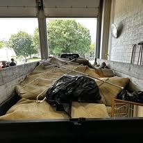 A bin of burlap sacks with a black trash bag inside, viewed from a doorway with a tree-lined outdoor view.