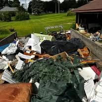 Dumpster filled with trash, including bags, wood, and debris, outdoors near a building and grass.