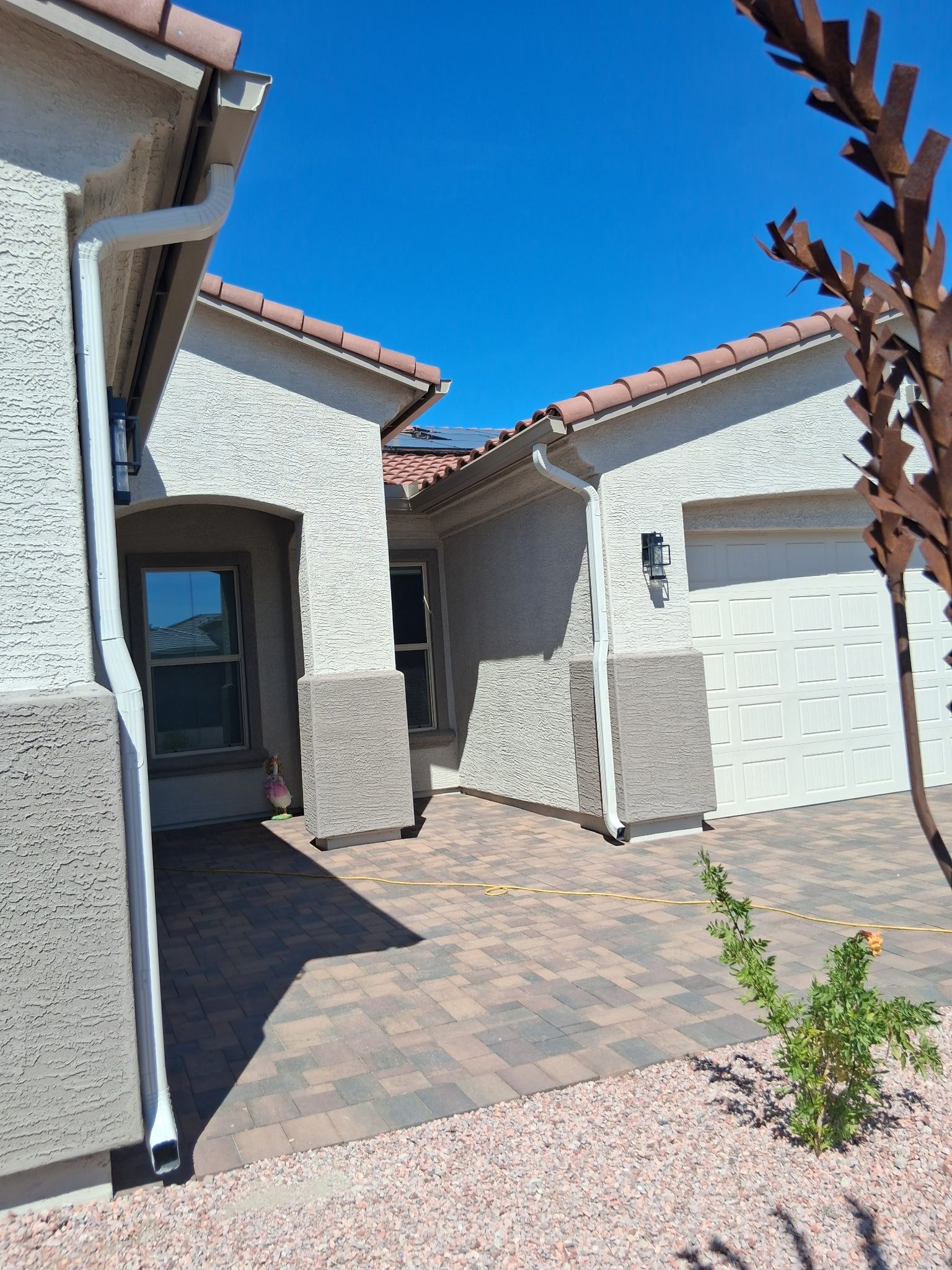 The exterior front of a light-stucco house with a paved driveway, tiled roof, and a small desert-landscaped yard.