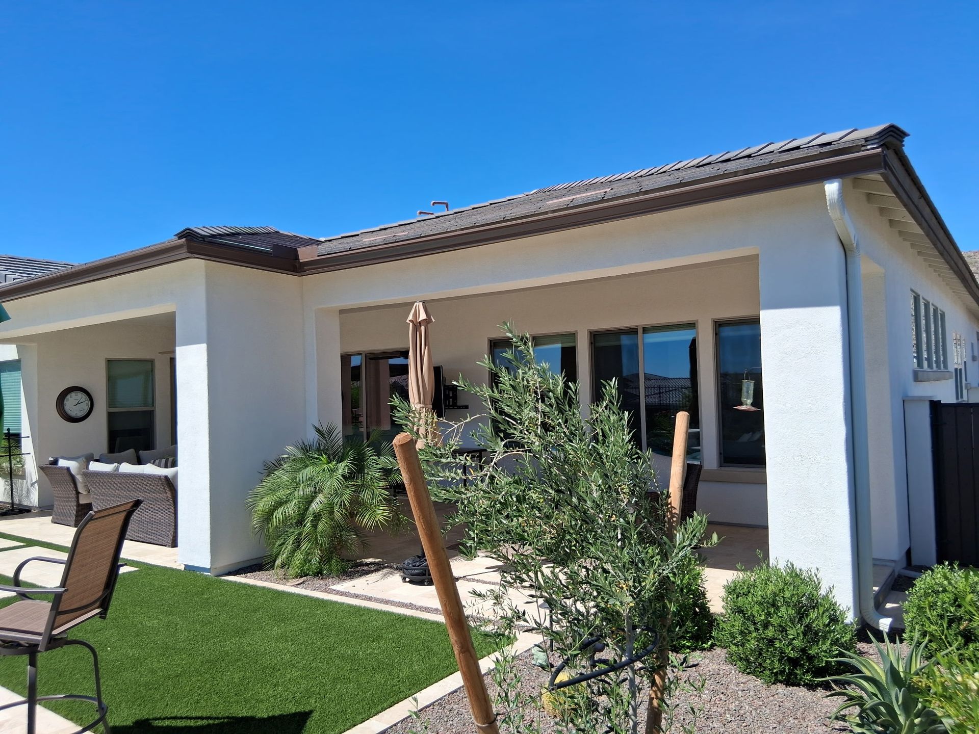 A covered patio area of a white house with a brown tile roof, outdoor furniture, green lawn, and desert landscaping.