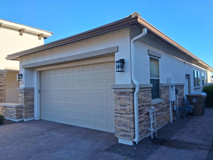 A beige garage with stone siding and a dark roof, adjacent to a sidewalk under a clear blue sky.