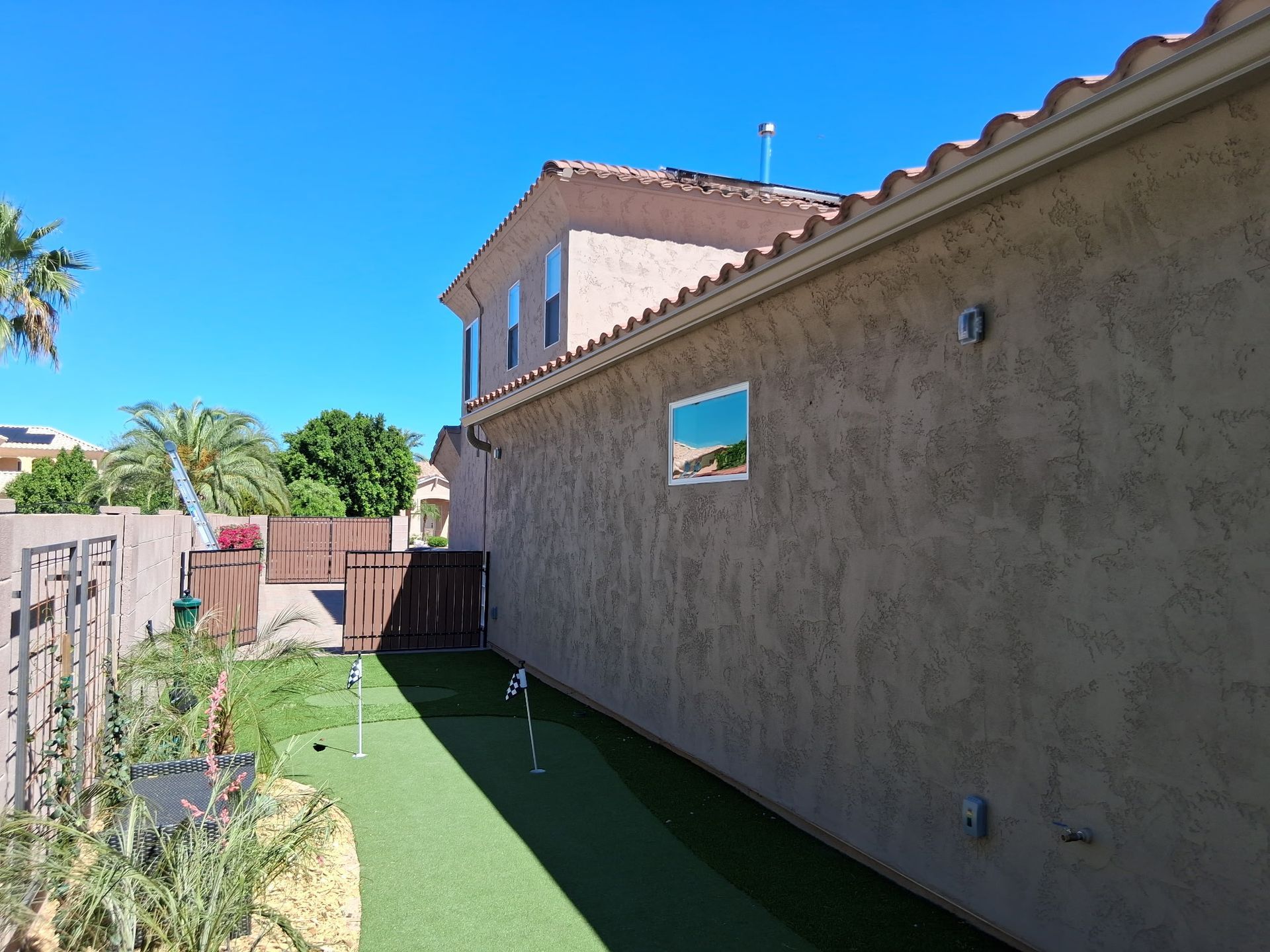 A side view of a house exterior with a small artificial putting green featuring two flags on a narrow side yard.