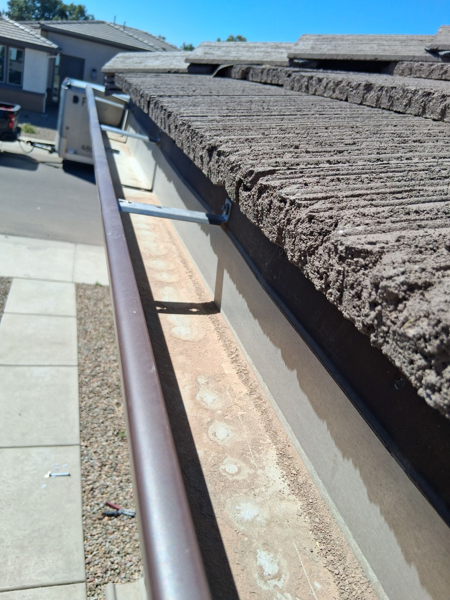 A metal gutter filled with light-colored debris, mounted beneath a brown textured roof on a sunny day.