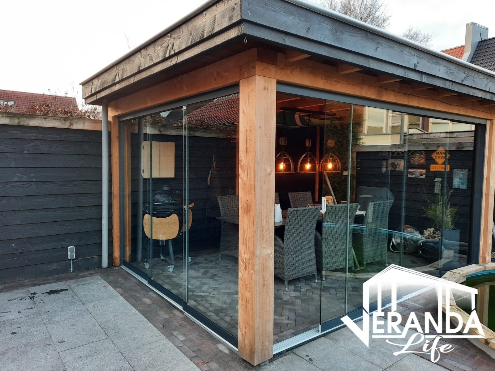 Een houten tuinhuisje met glazen schuifdeuren en een eettafel en stoelen.