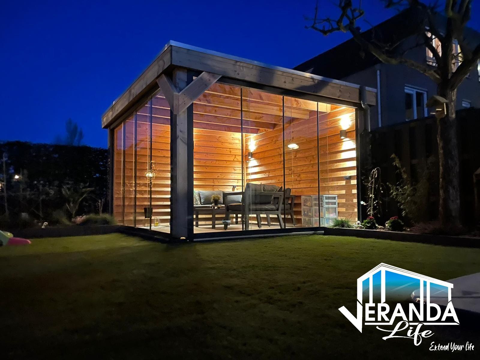 Een houten tuinhuisje met veel glazen ramen wordt 's avonds verlicht.