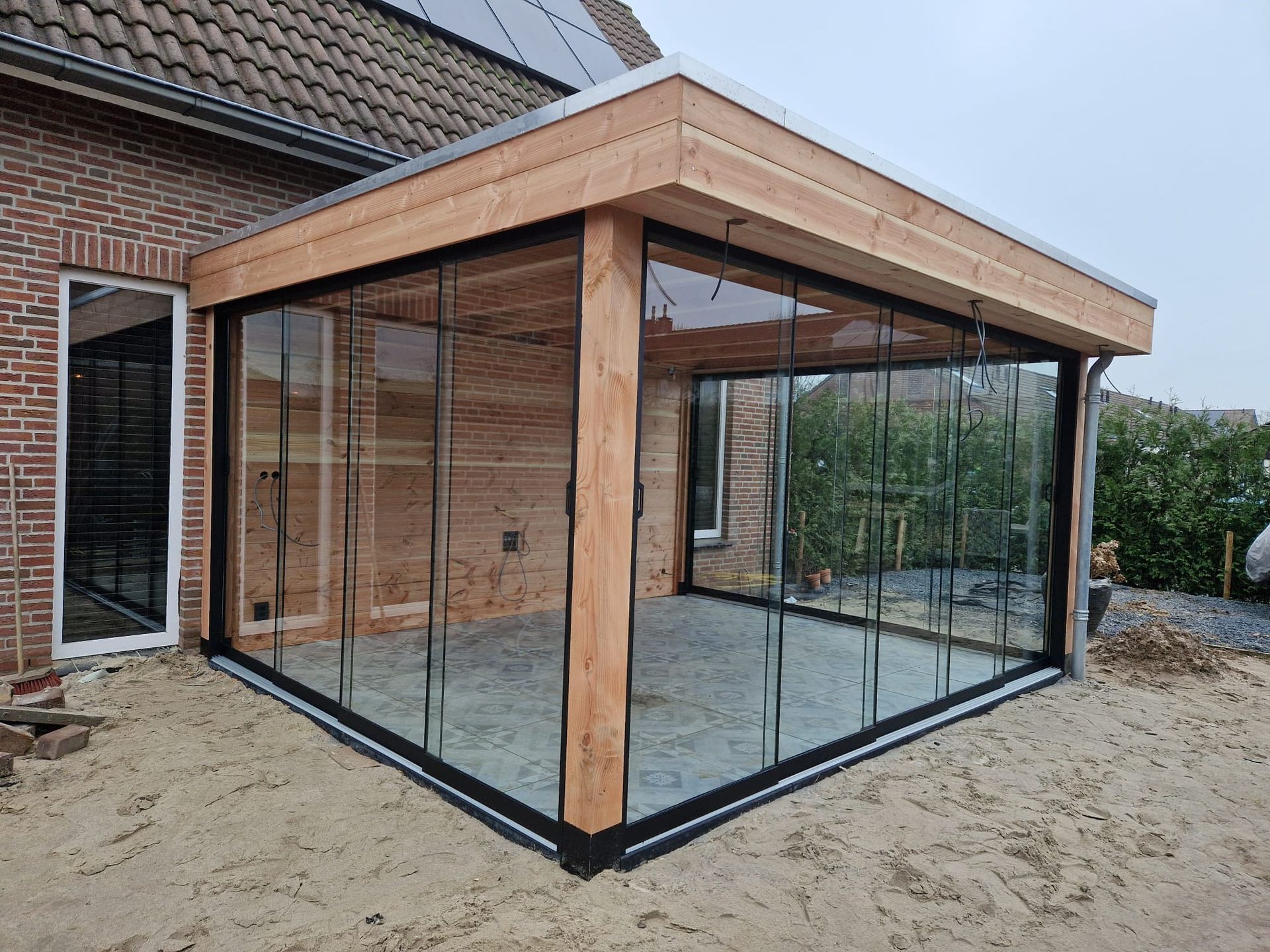 Een houten tuinhuisje met glazen schuifdeuren en een bakstenen gebouw op de achtergrond.